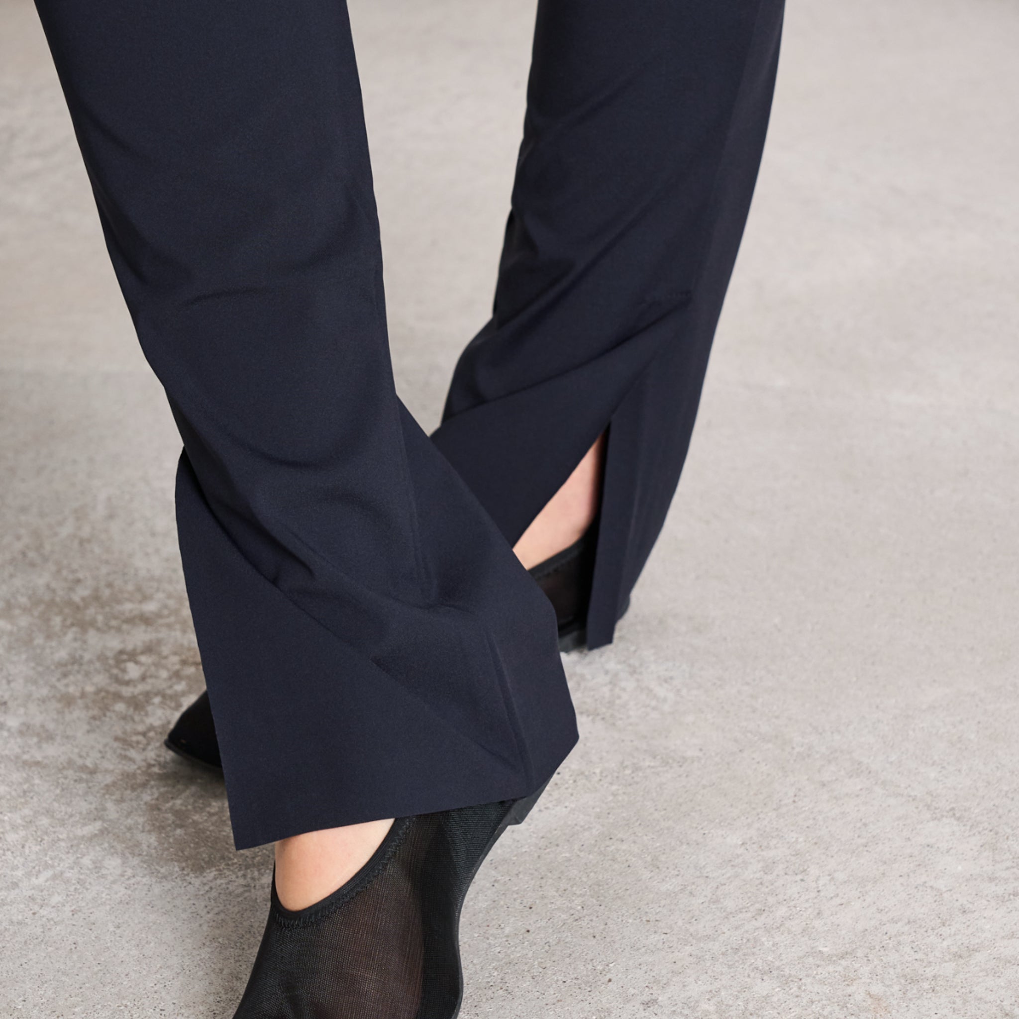 Nahaufnahme von schwarzen Stoffhosen mit seitlichen Schlitzen über schwarzen Halbschuhen auf einem grauen Boden.