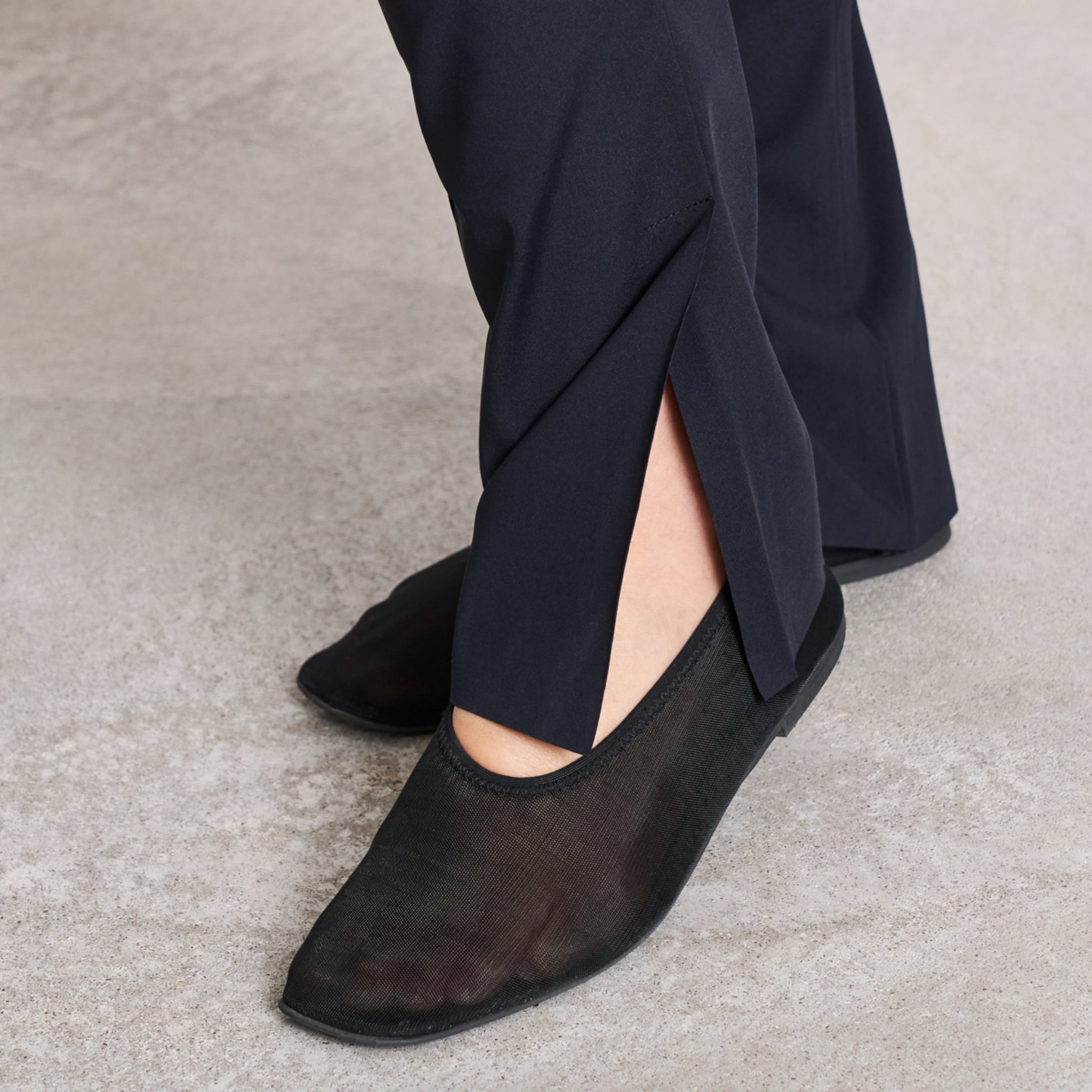 Schwarze Schuhe aus Netzstoff auf Betonboden, getragen zu einer Hose mit seitlichem Schlitz am Bein.