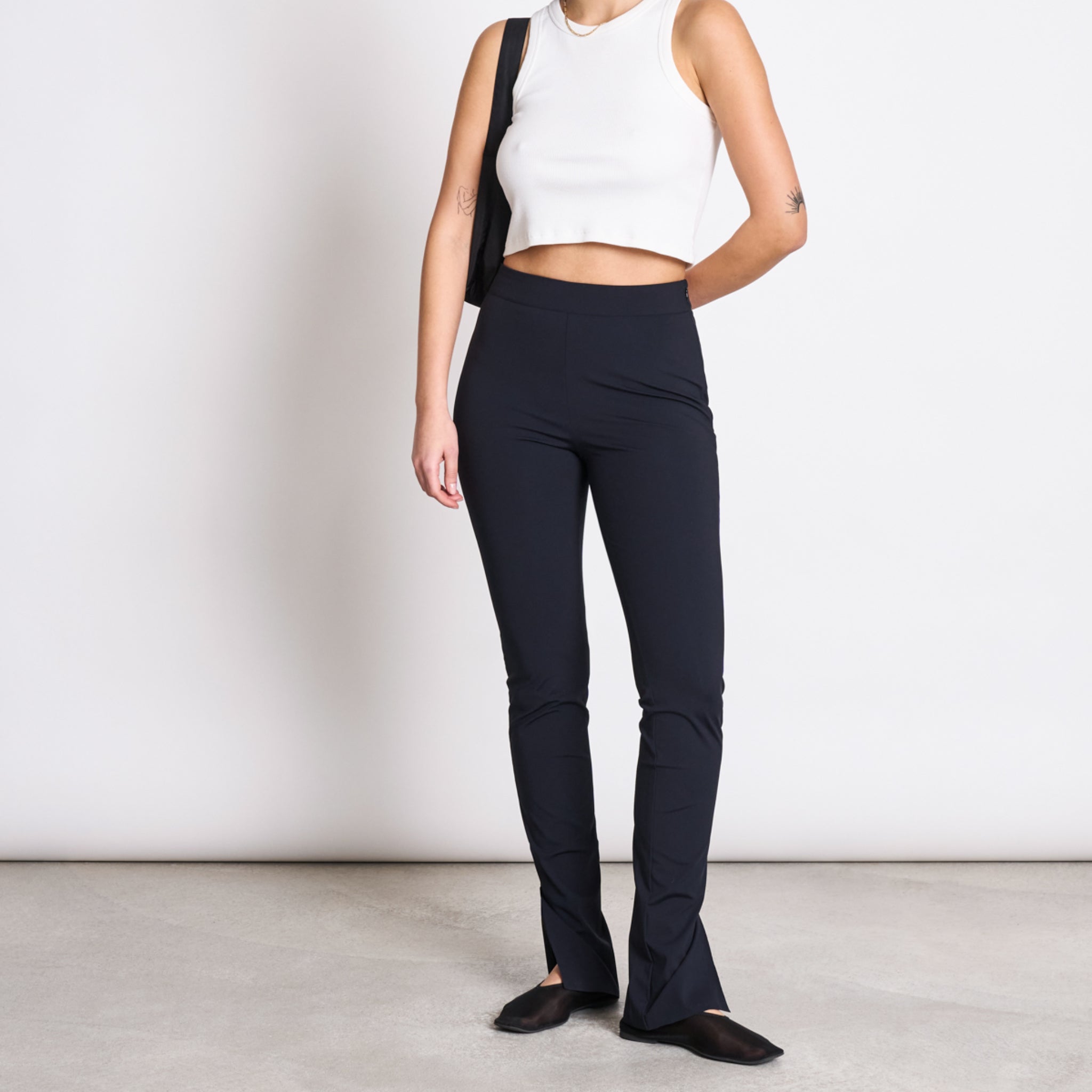 Person in weißem Crop-Top und schwarzer Hose steht vor einer weißen Wand auf grauem Boden.