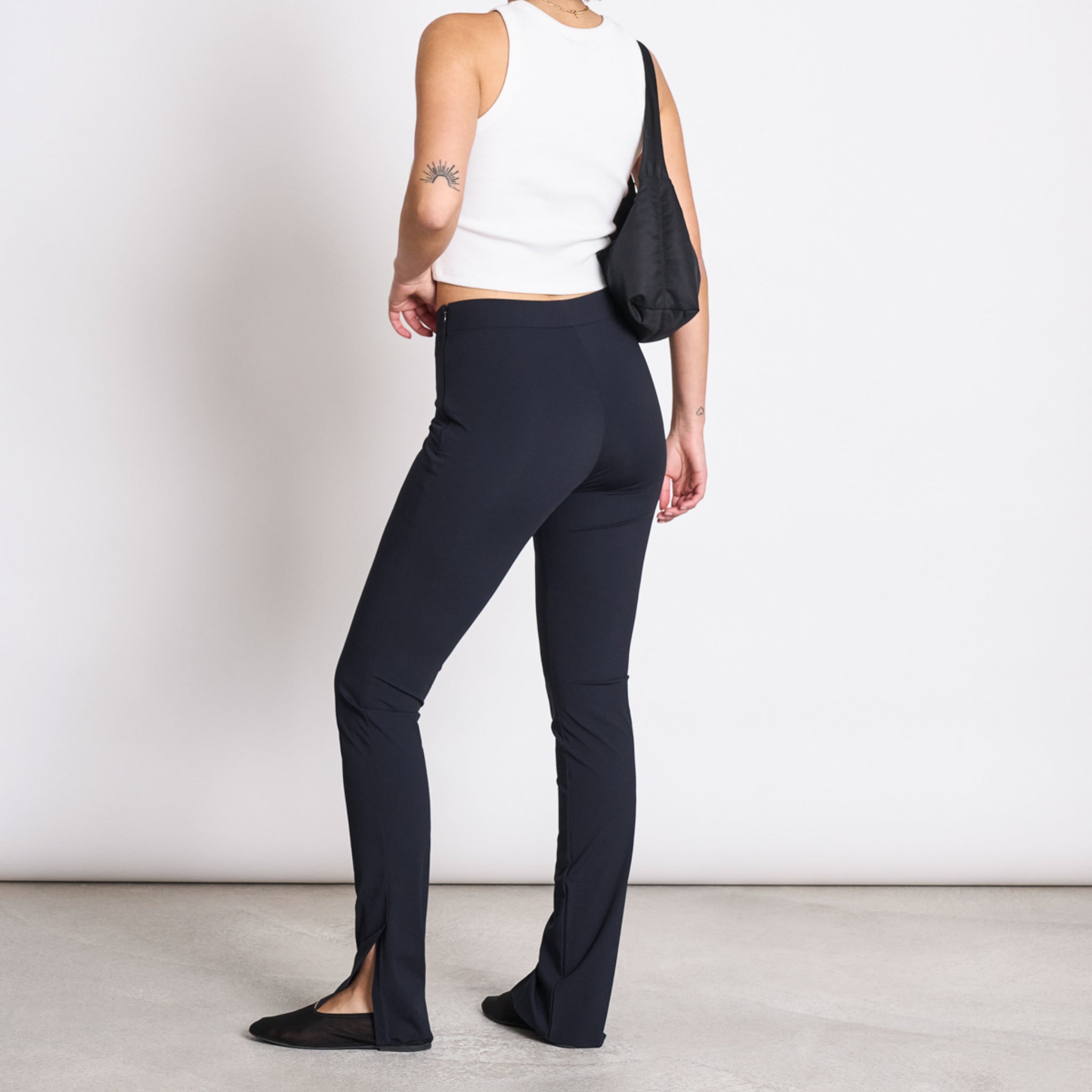 Person in weißem Top und schwarzer Hose steht mit Tasche über der Schulter; Seitenansicht.