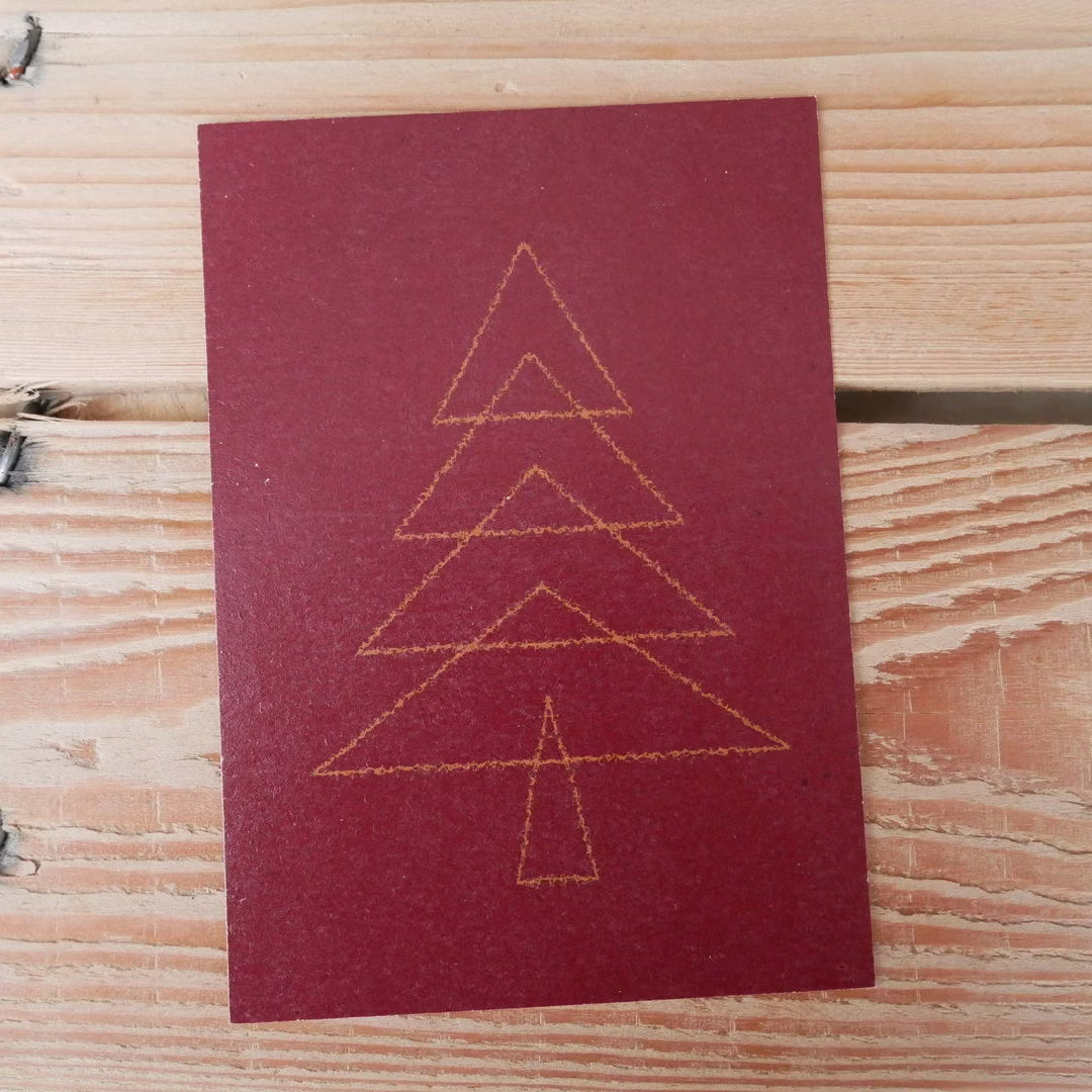 Rote Grußkarte mit stilisiertem Weihnachtsbaum aus goldenen Linien, auf einem Holztisch liegend.