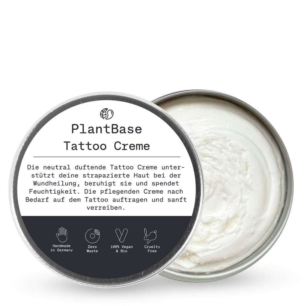 Eine runde Dose PlantBase Tattoo Creme mit teilweise geöffnetem Deckel, der eine weiße Creme im Inneren offenbart. Das Etikett ist auf Deutsch und enthält Symbole für Handmade, Zero Waste, Vegan und Cruelty Free.