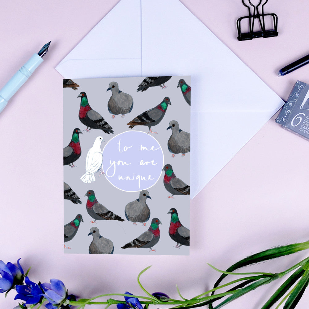 Eine Grußkarte mit illustrierten Tauben und einer weißen Taube, mit dem handgeschriebenen Text "To me you are unique". Die Karte befindet sich in einem Umschlag, umgeben von einem Stift, einer Büroklammer und blauen Blumen.