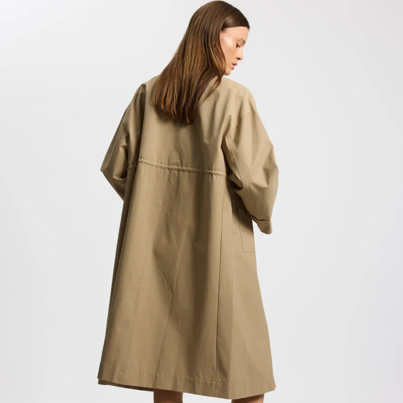 Eine Frau mit glattem braunem Haar, die einen locker sitzenden beigen Trenchcoat trägt, steht mit dem Rücken zur Kamera vor einem schlichten hellen Hintergrund.