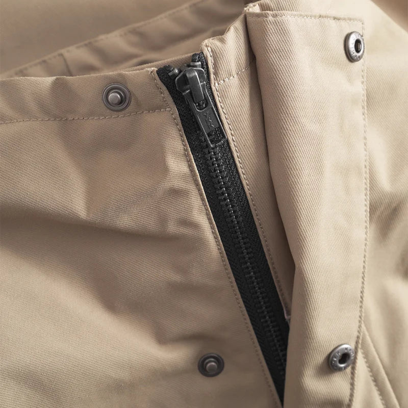 Nahaufnahme einer beigen Stoffjacke mit schwarzem Reißverschluss und Druckknöpfen an einer Klappe, die die Details des Verschlusses des Kleidungsstücks hervorheben.