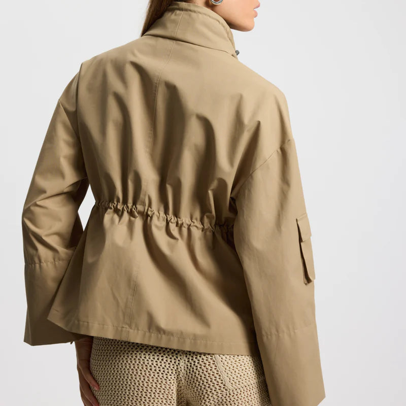 Eine Frau steht mit dem Gesicht zur Wand und trägt eine beigefarbene, taillierte Jacke und beigefarbene Strickshorts. Sie hat glattes braunes Haar und trägt Ohrringe vor einem schlichten Hintergrund.