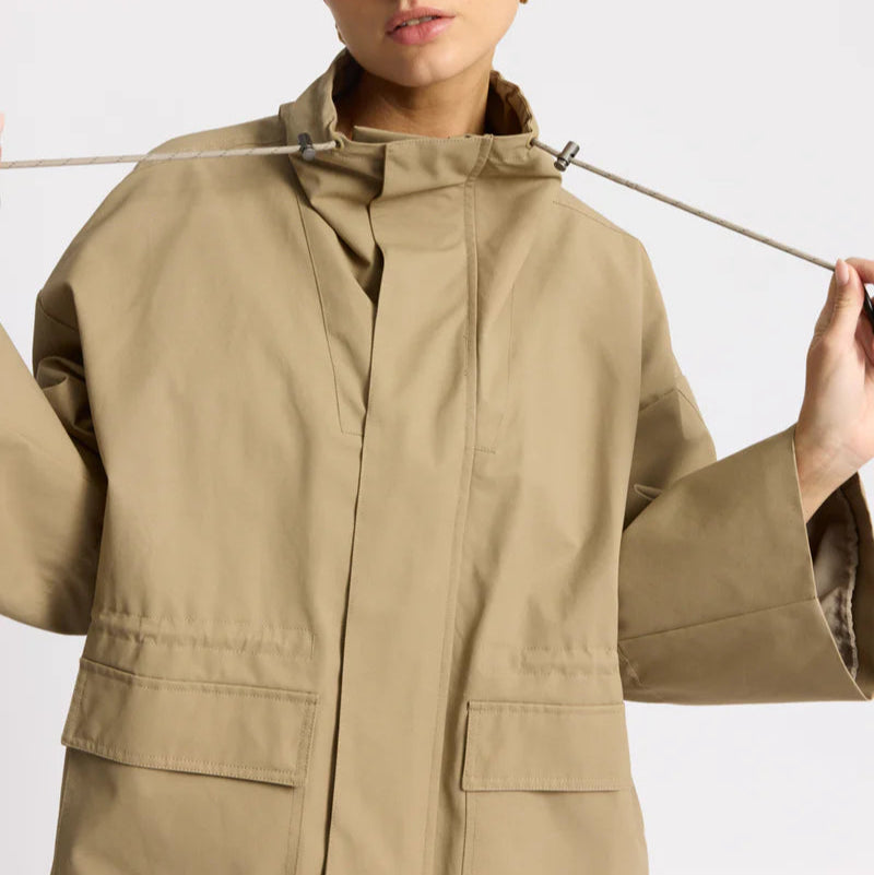 Eine Frau, die eine locker sitzende, hellbraune Jacke mit großen Vordertaschen trägt, zieht in der Nähe des Kragens an der Kordel der Jacke und blickt vor einem schlichten hellen Hintergrund zur Seite.