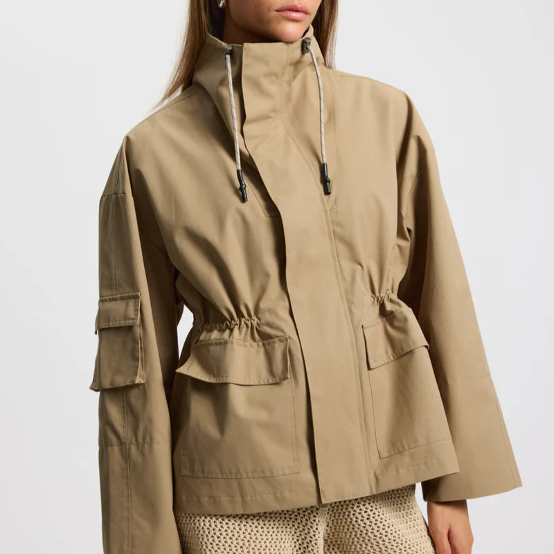 Eine Frau trägt eine beigefarbene, hochgeschlossene Funktionsjacke mit Kordelzug und Pattentaschen auf der Vorderseite, gepaart mit hellen, strukturierten Shorts, und steht vor einem einfarbigen weißen Hintergrund.