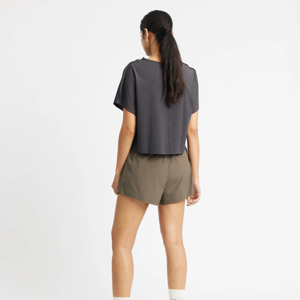 Frau in dunkelblauem T-Shirt und olivgrünen Shorts steht mit dem Rücken zur Kamera vor weißem Hintergrund.