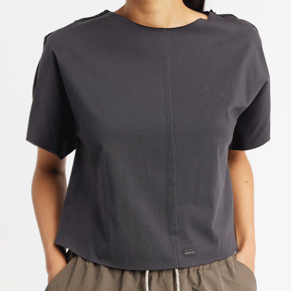 Person trägt ein dunkles T-Shirt mit kurzen Ärmeln und eine beige Hose, Hände in den Hosentaschen.