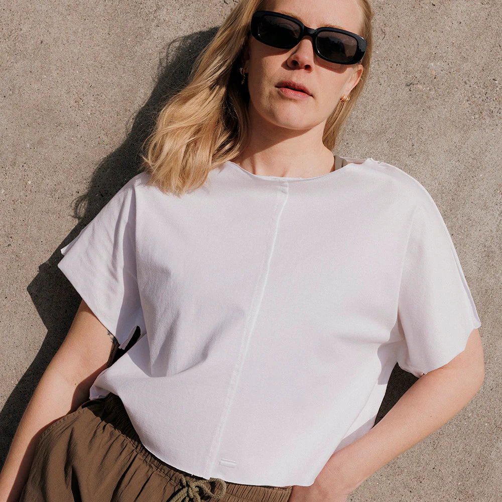 Frau mit Sonnenbrille trägt weißes T-Shirt und braune Hose, lehnt sich gegen eine Betonwand.