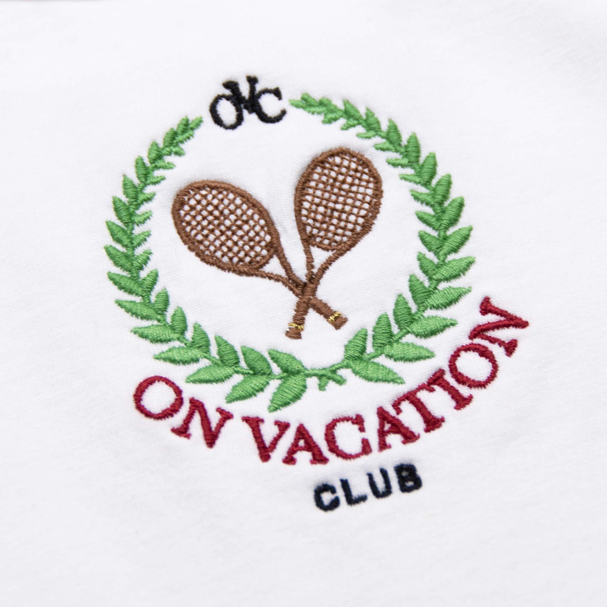 Stickerei von zwei Tennisschlägern in einem grünen Lorbeerkranz mit dem Text "ON VACATION CLUB" darunter auf Stoff.