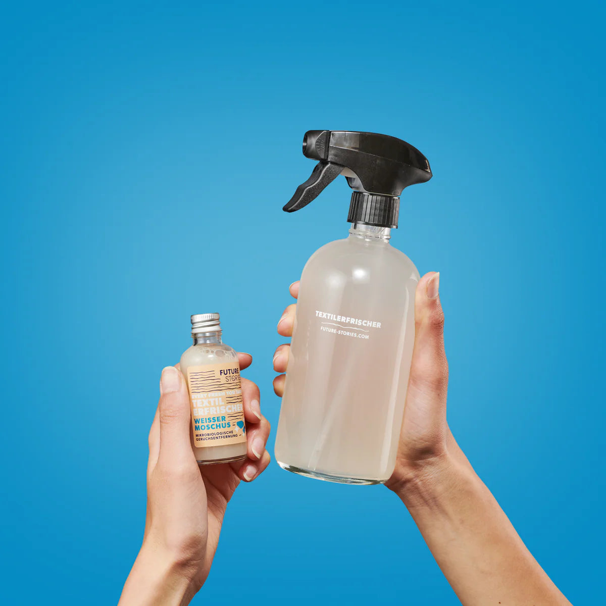 Zwei Hände halten Flaschen vor einem blauen Hintergrund: eine Hand hält eine kleine Nachfüllflasche, die andere eine größere Sprühflasche mit der Aufschrift TEXTILERFRISCHER.