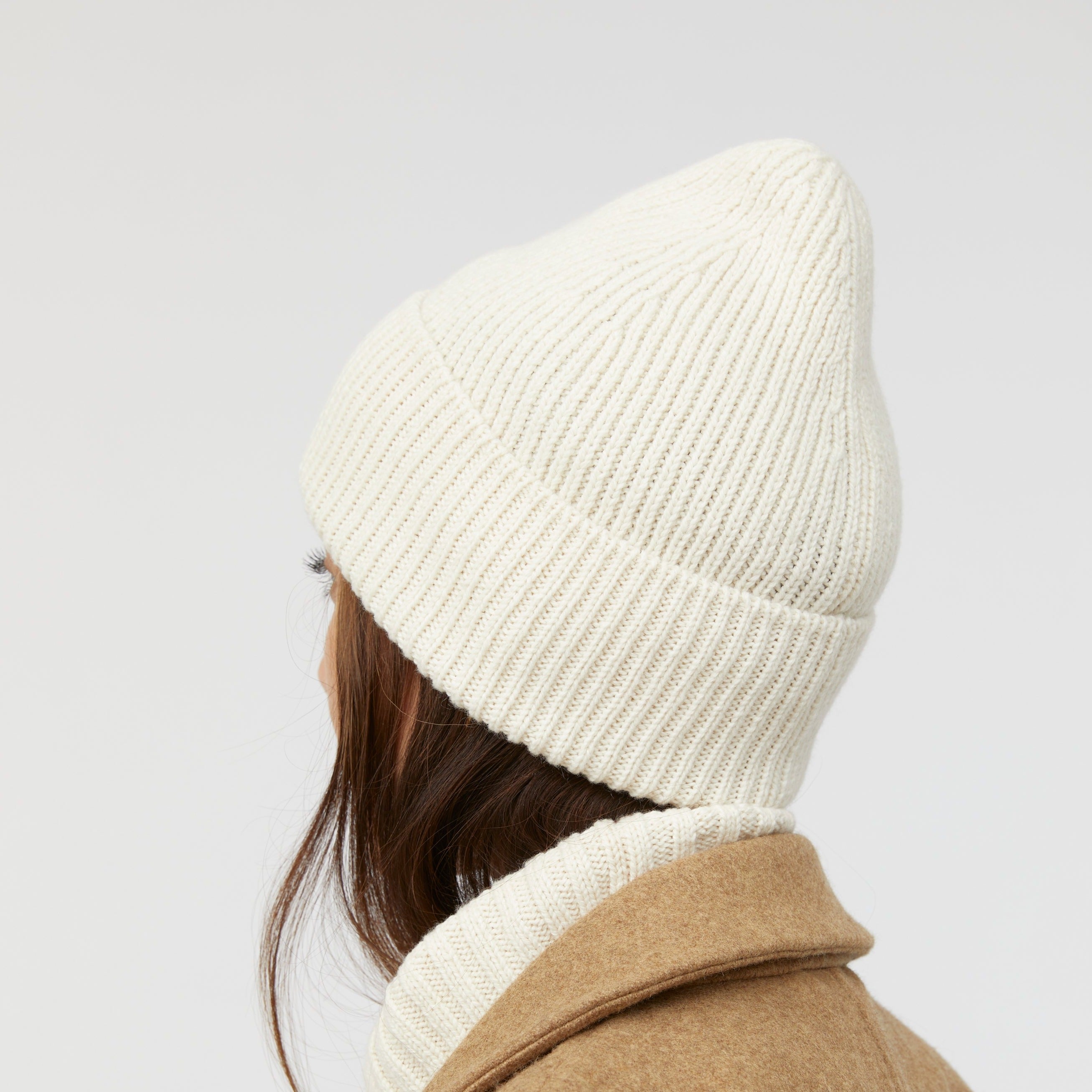 Eine Person trägt eine cremefarbene Strickmütze und einen braunen Mantel, von hinten fotografiert.