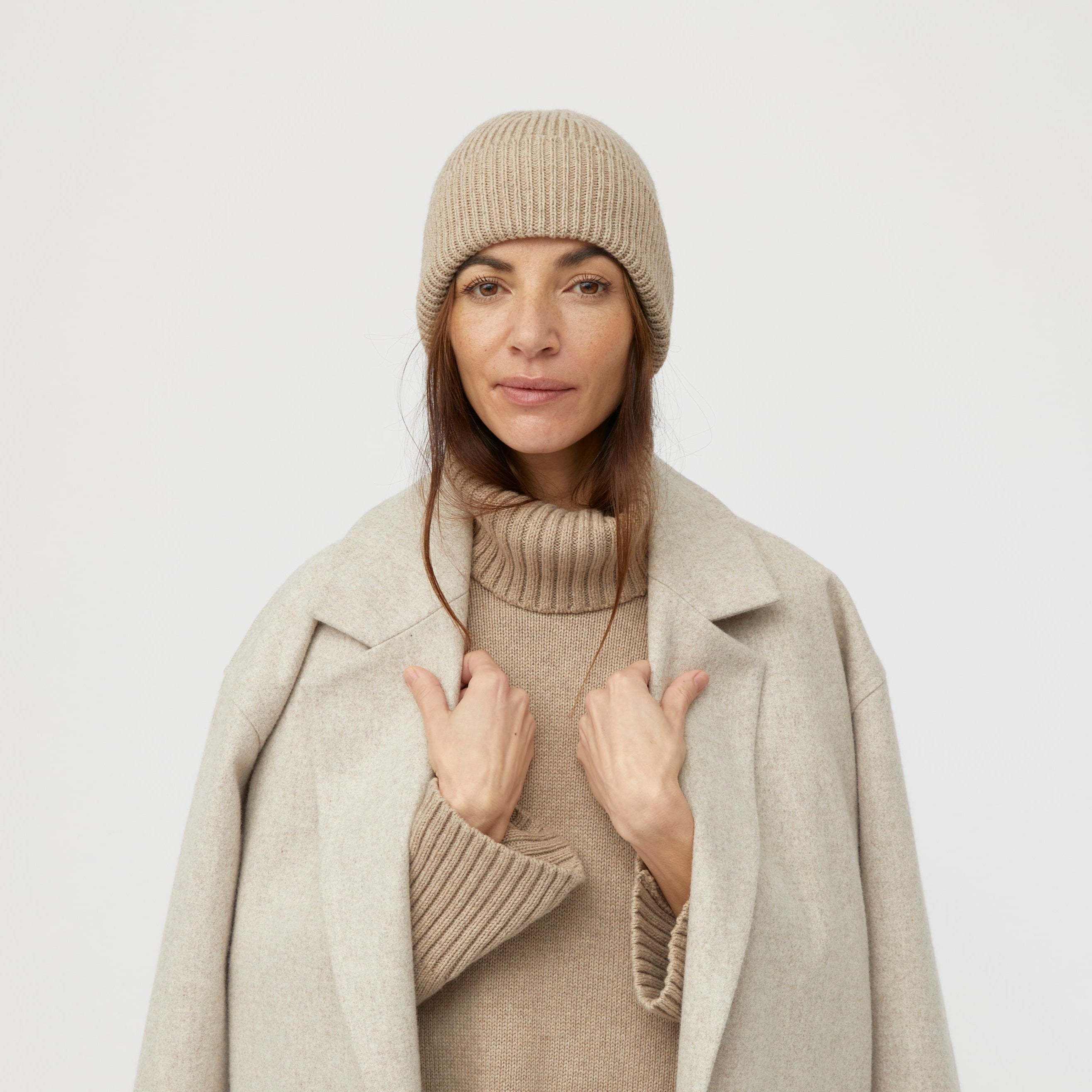 Frau in heller Winterkleidung, mit gestrickter Mütze und Mantel, steht vor weißem Hintergrund.