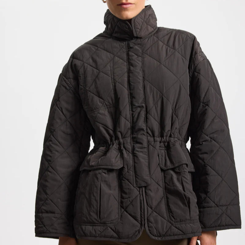 Eine Person trägt eine schwarze, gesteppte Jacke mit hohem Kragen, Frontverschluss und großen Vordertaschen. Die Jacke hat einen Taillengürtel und der Hintergrund ist einfarbig weiß. Das Gesicht der Person ist teilweise außerhalb des Rahmens.
