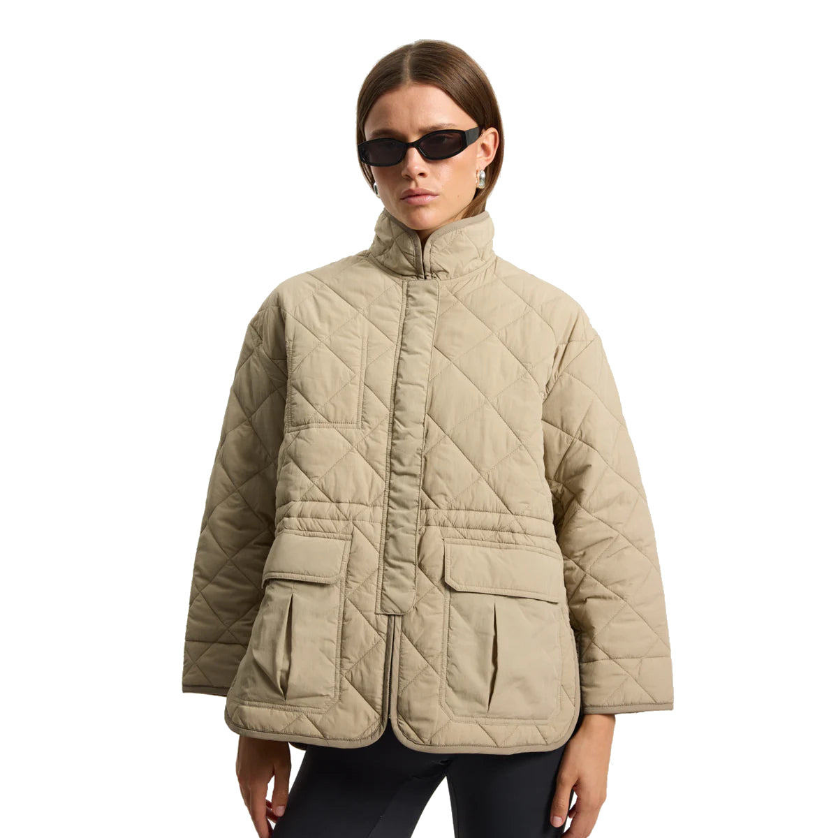 Eine Frau mit einer schwarzen Sonnenbrille und einer beigen Steppjacke mit großen Vordertaschen steht vor einem schlichten weißen Hintergrund.
