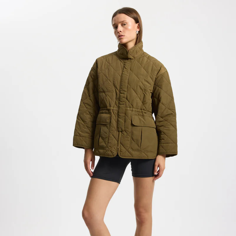 Eine Frau steht in einer olivgrünen Steppjacke mit großen Vordertaschen und schwarzen, eng anliegenden Shorts vor einem schlichten weißen Hintergrund.