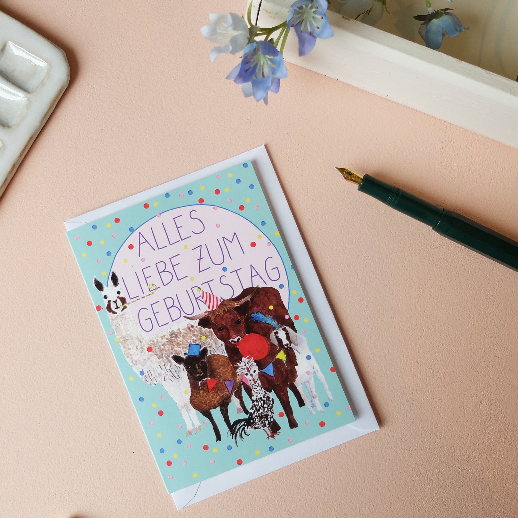 Geburtstagskarte mit einem Lama, einem Stier, zwei Schafen und einem Hahn, umgeben von bunten Punkten, auf einem Tisch.