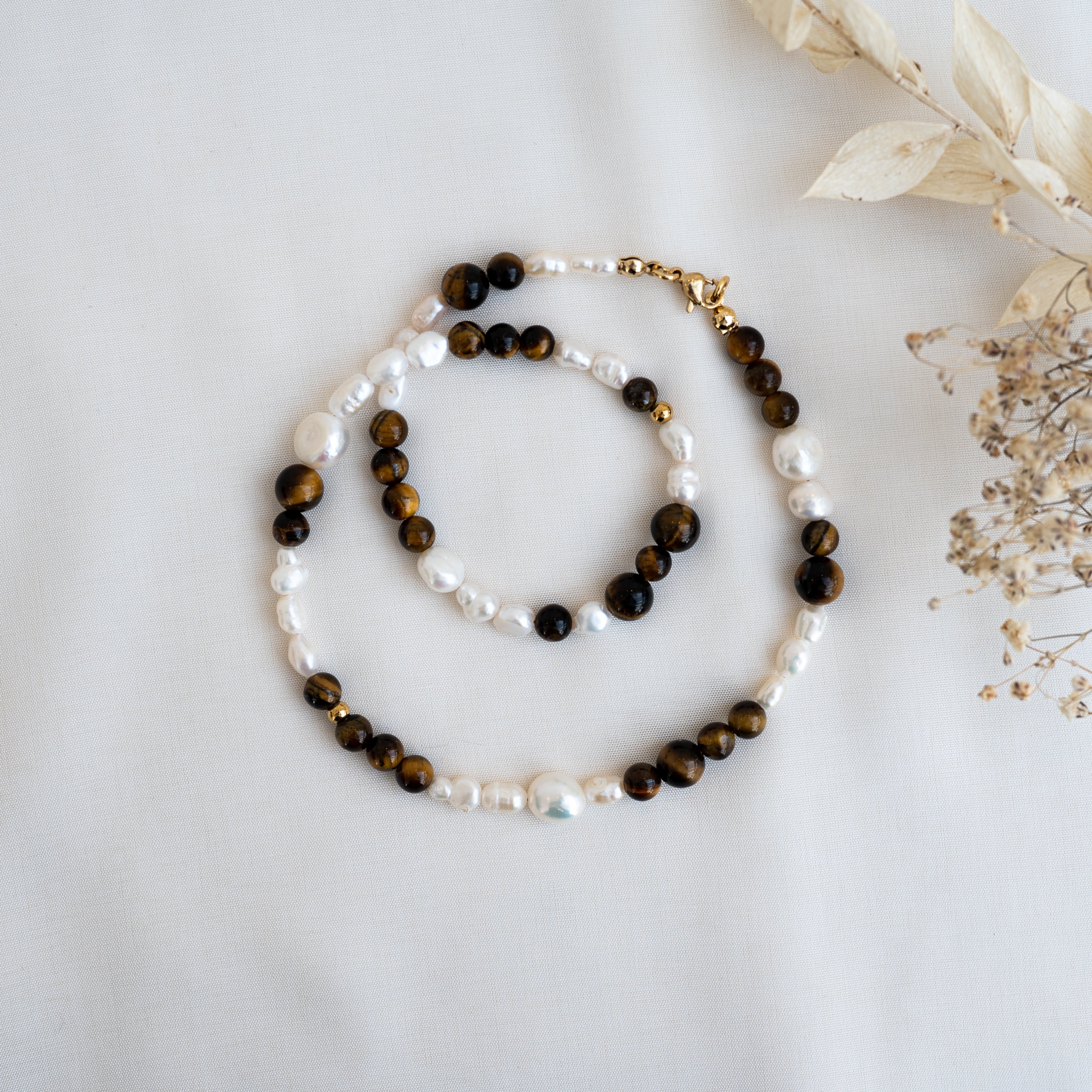 Eine Perlenkette mit abwechselnd braunen Tigeraugensteinen und weißen Perlen ist auf einem weißen Stoffhintergrund neben getrockneten beigen Blumen ausgestellt.