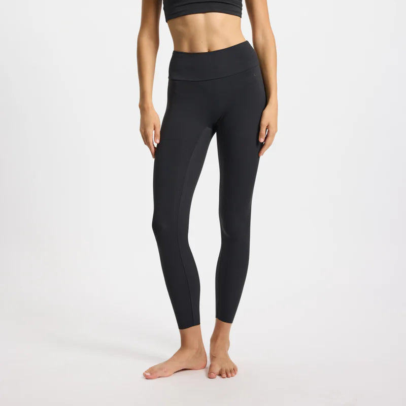 Eine Frau, die einen schwarzen Sport-BH und schwarze Leggings mit hoher Taille trägt, steht barfuß auf einem einfarbig weißen Hintergrund, die Arme locker an den Seiten. Ihr Gesicht ist nicht zu sehen.