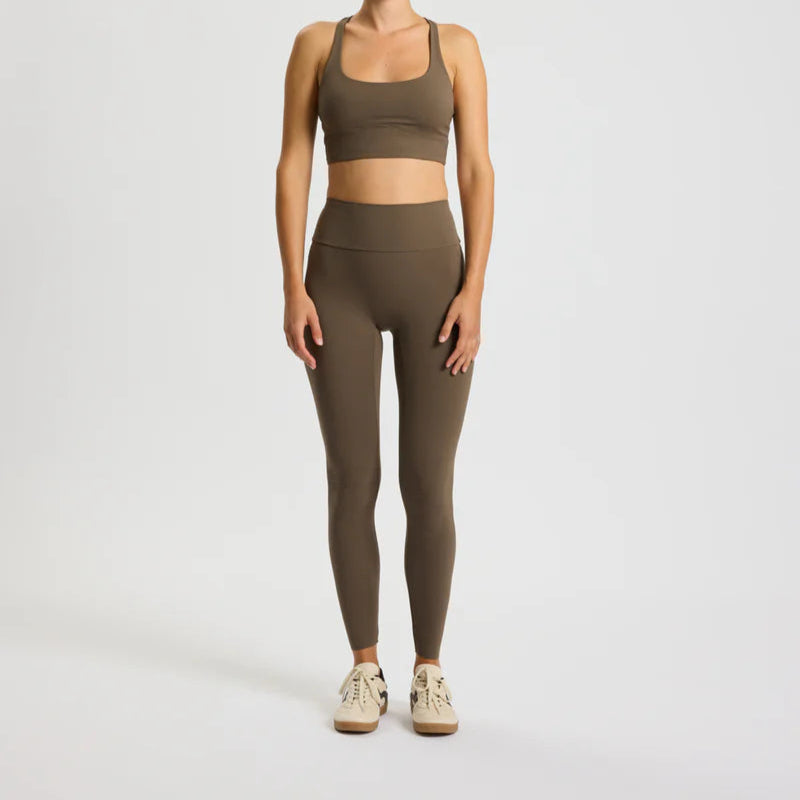 Eine Person steht mit dem Gesicht nach vorne und trägt einen braunen Sport-BH und hoch taillierte Leggings sowie beigefarbene Turnschuhe vor einem schlichten weißen Hintergrund.