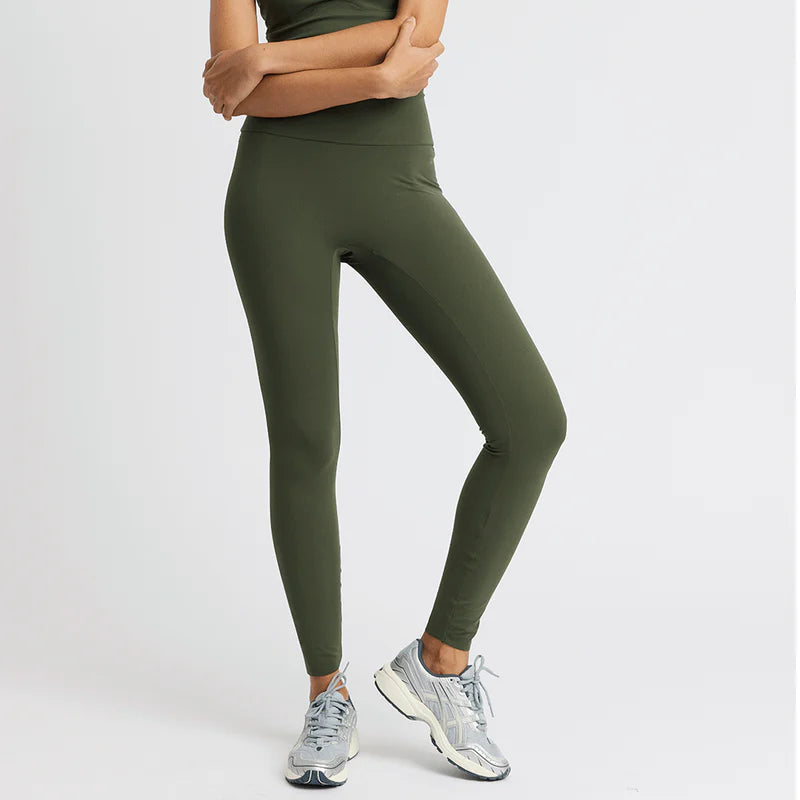 Eine Frau steht vor einem schlichten Hintergrund und trägt ein olivgrünes Tanktop und dazu passende Leggings mit hoher Taille, gepaart mit grauen und weißen Turnschuhen. Ihre Arme sind verschränkt und sie blickt selbstbewusst in die Kamera.