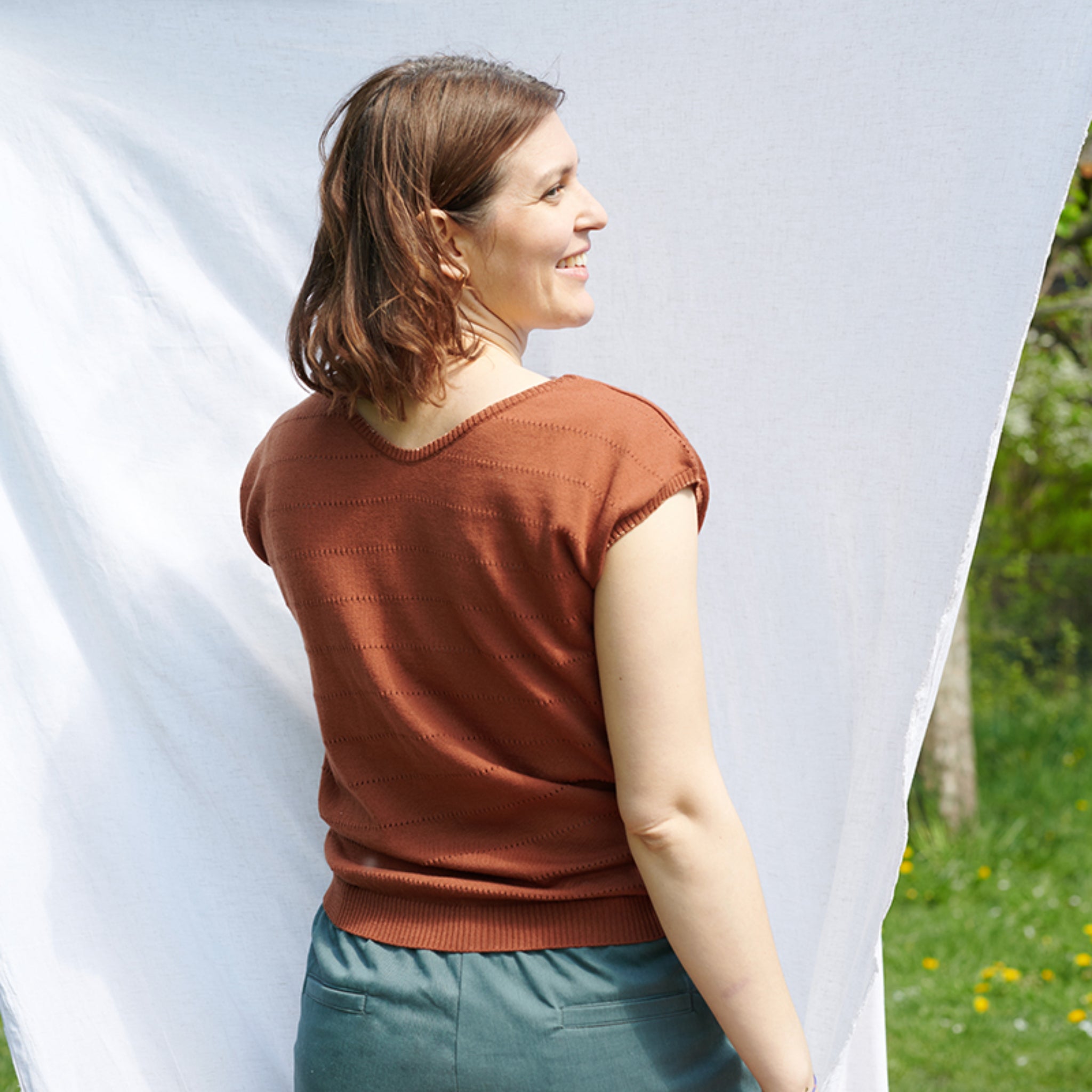 Frau mit braunem Top steht draußen vor einem weißen Tuch und lächelt.