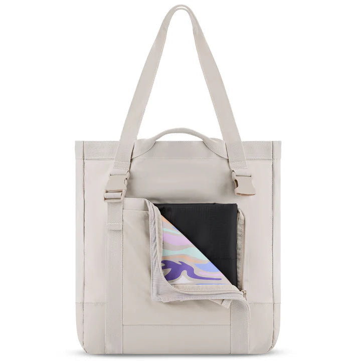 Eine beigefarbene Tragetasche mit Schulterriemen wird mit geöffneter Vordertasche gezeigt, in der ein schwarzer Beutel und ein bunter, wellenförmig gemusterter Gegenstand zu sehen sind. Die Tasche hat ein minimalistisches und modernes Design.