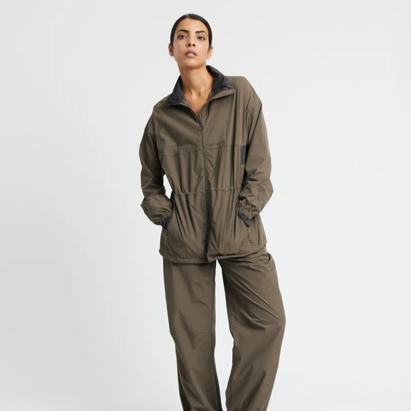 Eine Person, die eine locker sitzende, olivgrüne Jacke und eine dazu passende Hose trägt, posiert vor einem schlichten weißen Hintergrund mit den Händen in den Jackentaschen.