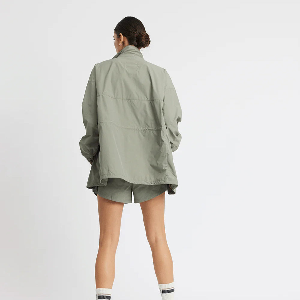 Person von hinten in olivgrüner Jacke und Shorts, auf neutralem Hintergrund stehend.