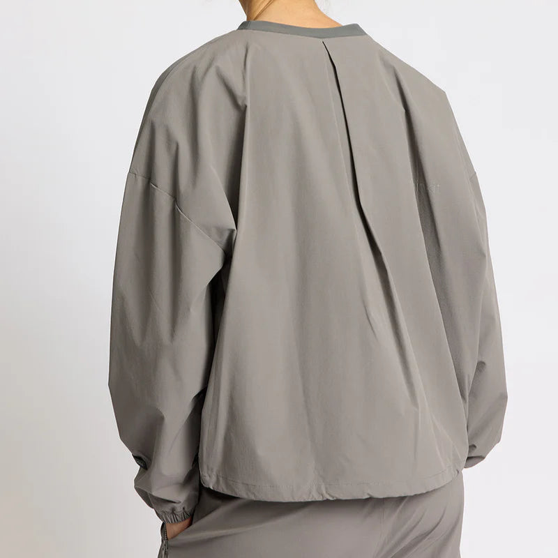 Eine Person, die eine lockere, langärmelige, hellgraue Jacke und eine dazu passende Hose trägt, wird von hinten vor einem schlichten, hellen Hintergrund gezeigt.