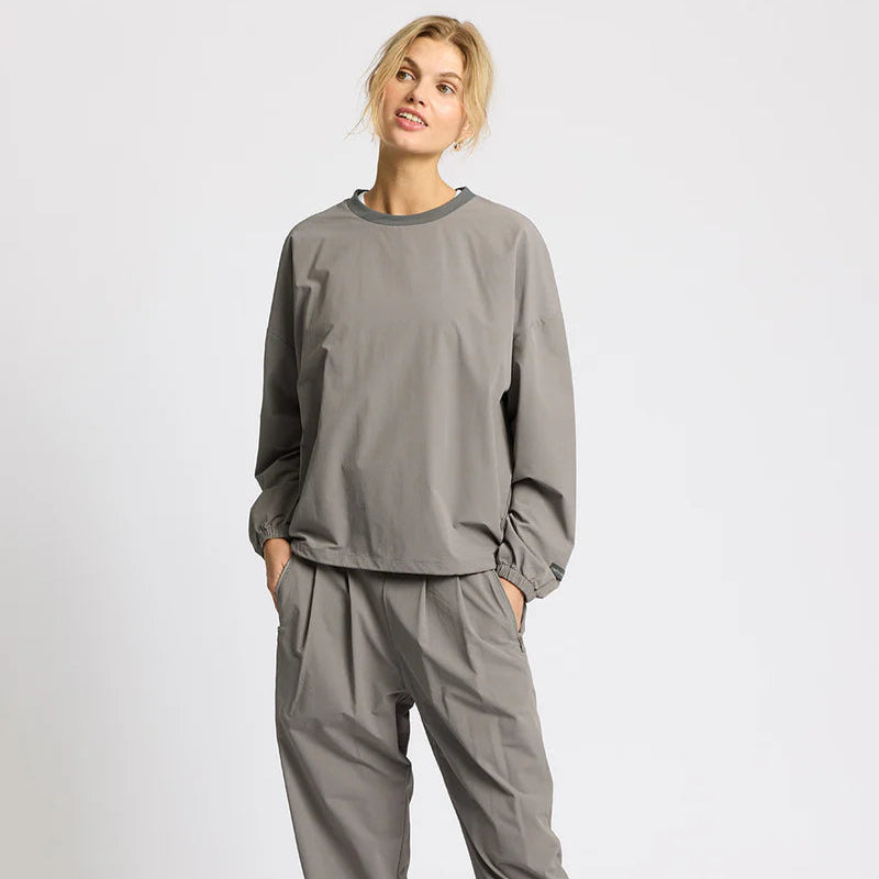 Eine Person steht vor einem schlichten weißen Hintergrund, trägt ein locker sitzendes, langärmeliges graues Oberteil und eine dazu passende graue Hose, hat die Hände in den Taschen und blickt mit entspannter Miene leicht zur Seite.