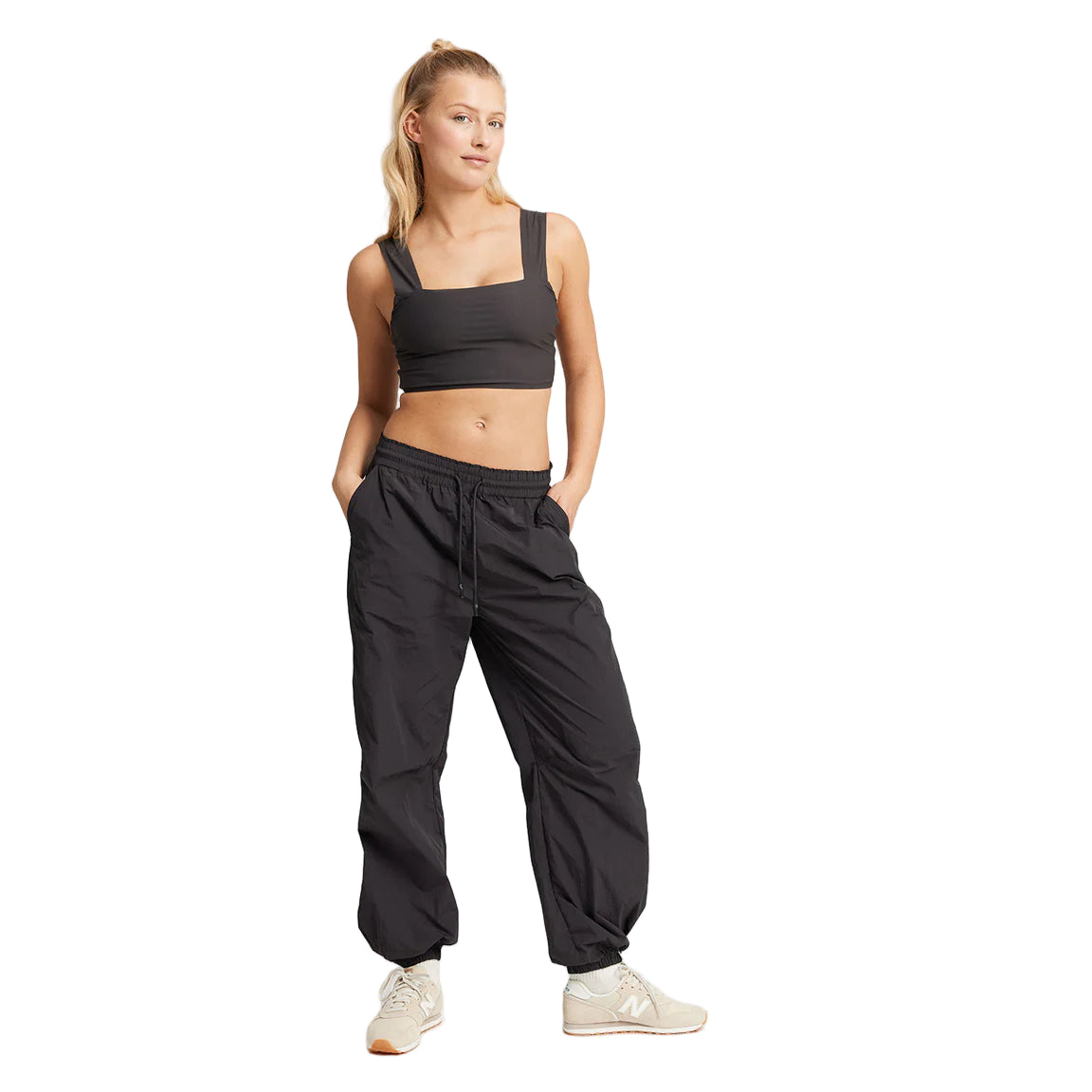 Eine Frau mit blondem Haar in einem Pferdeschwanz trägt einen schwarzen Sport-BH, eine schwarze Jogginghose und beigefarbene Turnschuhe. Sie steht mit leicht angewinkelten Händen in ihren Taschen vor einem weißen Hintergrund.