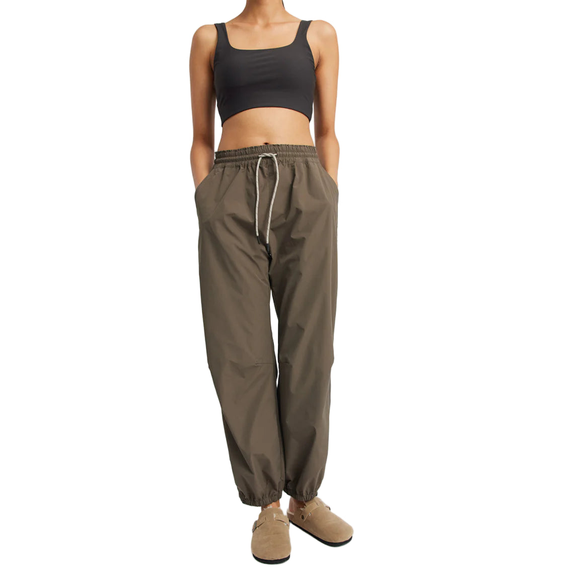 Eine Person, die ein schwarzes Crop-Top, eine olivgrüne Jogginghose mit Kordelzug in der Taille und hellbraune Slipper trägt, steht mit den Händen in den Taschen vor einem schlichten weißen Hintergrund.