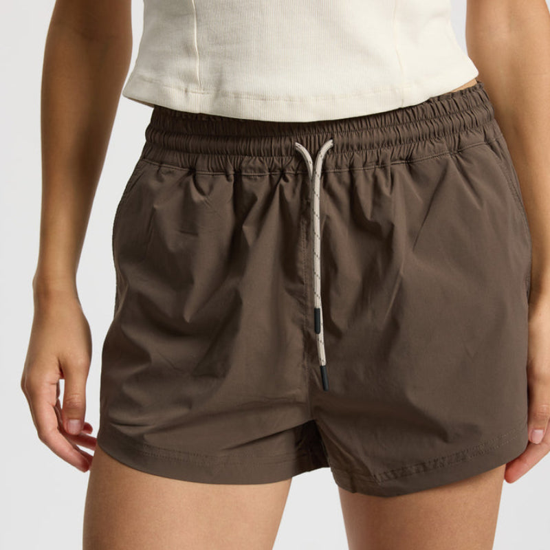 Eine Person, die ein weißes ärmelloses Oberteil und eine braune Hose mit Kordelzug trägt, steht vor einem unifarbenen Hintergrund und hält die Arme locker an der Seite.