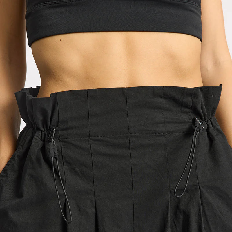 Nahaufnahme einer Person, die eine hochtaillierte schwarze Hose mit verstellbaren Kordelzügen und ein schwarzes Crop-Top trägt, wobei ihre Taille und die Hände in den Hosentaschen zu sehen sind.