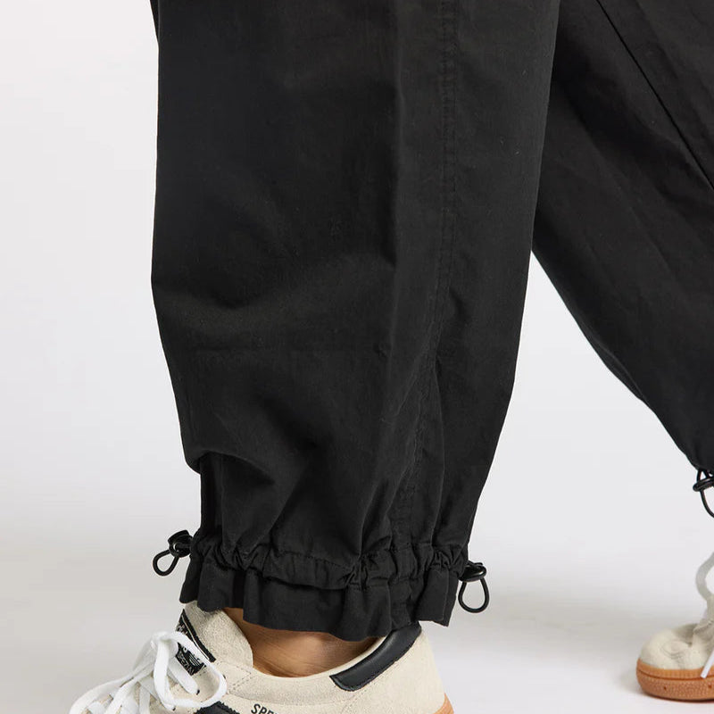 Nahaufnahme einer Person, die eine schwarze Hose mit Kordelzug und beigefarbene Turnschuhe mit schwarzen Streifen und Gummisohlen trägt und auf einer weißen Fläche steht.