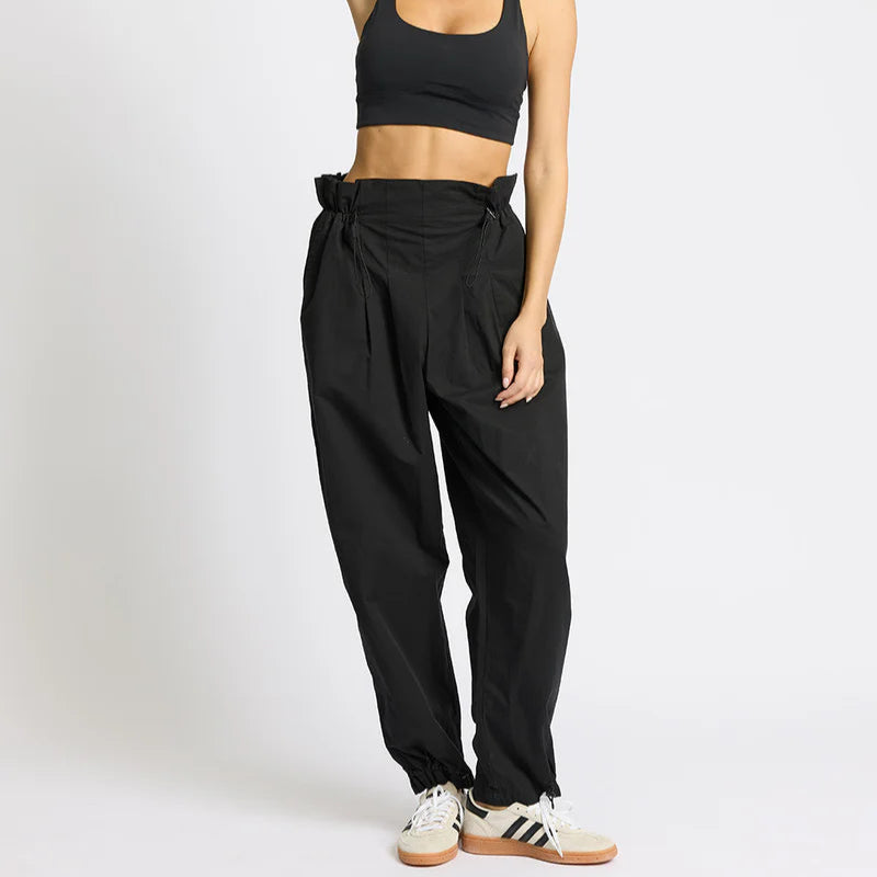 Eine Frau steht vor einem schlichten Hintergrund und trägt einen schwarzen Sport-BH, eine hochgeschnittene schwarze Paperbag-Hose und weiße Adidas-Turnschuhe. Sie hat einen Arm angewinkelt, die Hand hinter dem Kopf und schaut selbstbewusst in die Kamera.