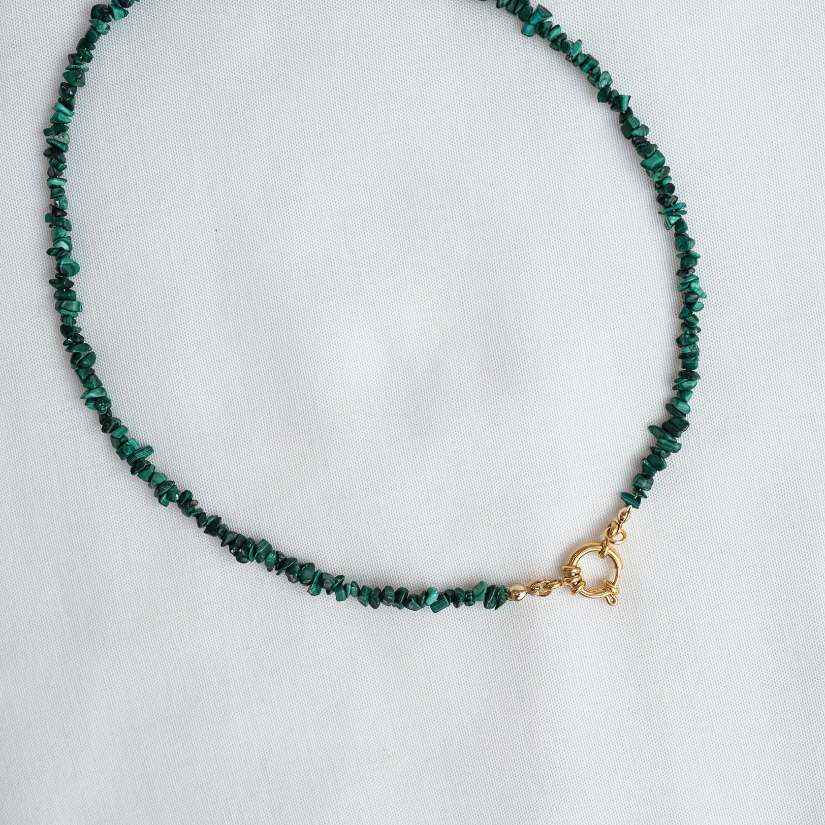 Eine Halskette aus kleinen, unregelmäßigen grünen Steinen mit einem goldenen Verschluss auf weißem Stoff.