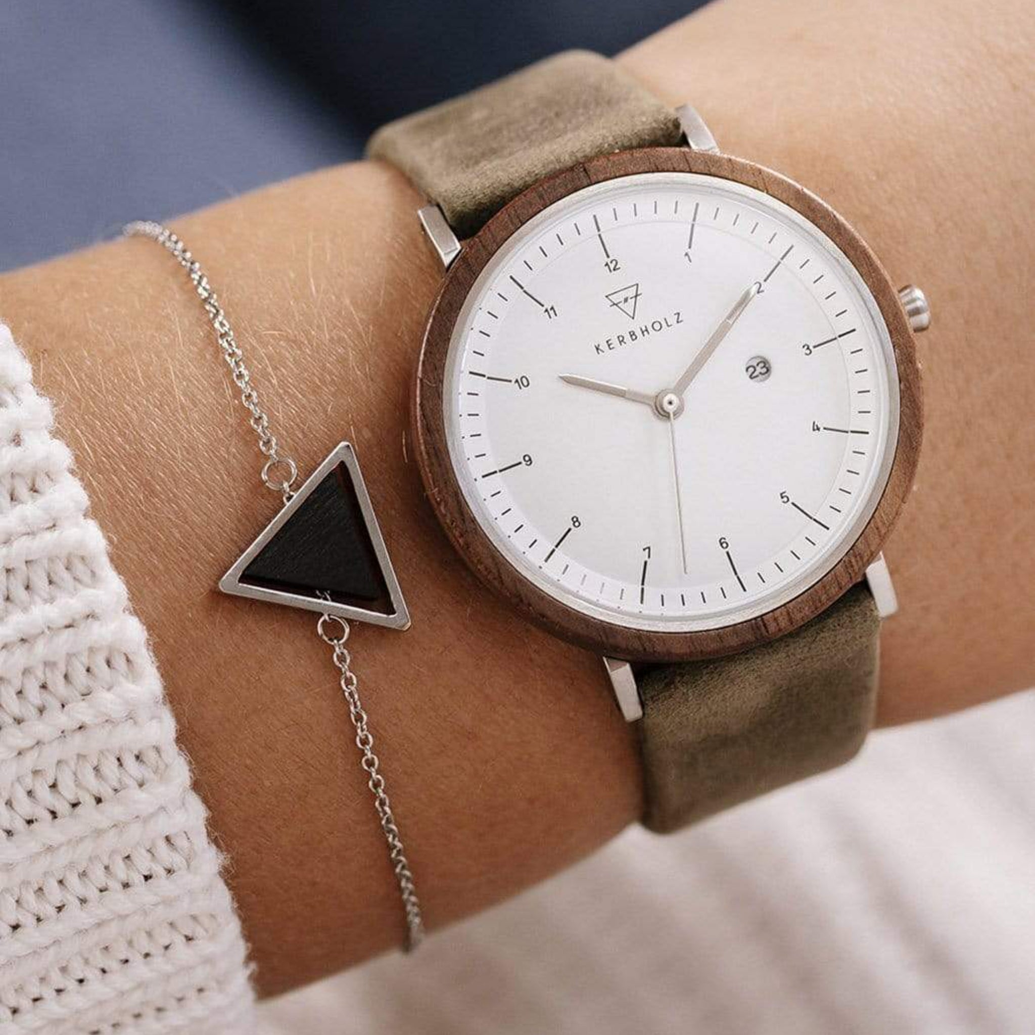 Arm mit einer Uhr mit weißem Zifferblatt und beigem Armband, daneben ein Armband mit dreieckigem Anhänger.