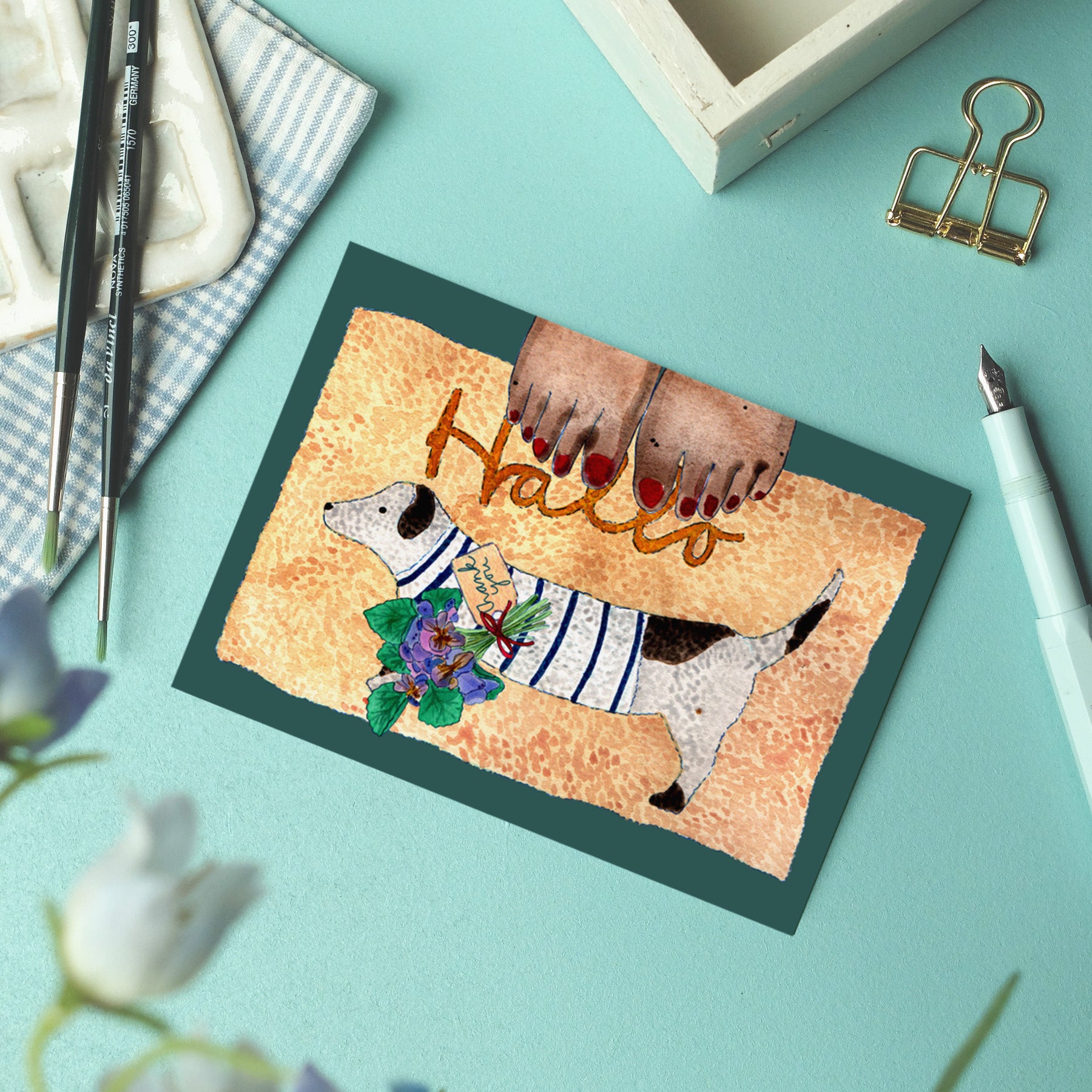 Eine Grußkarte mit Hallo zeigt einen Hund in einem gestreiften Hemd, der Blumen im Maul hält, und darüber ein Paar lackierte Zehennägel. Die Karte liegt auf einem Schreibtisch, daneben liegen ein Stift, Pinsel, eine Büroklammer und ein kariertes Tuch.