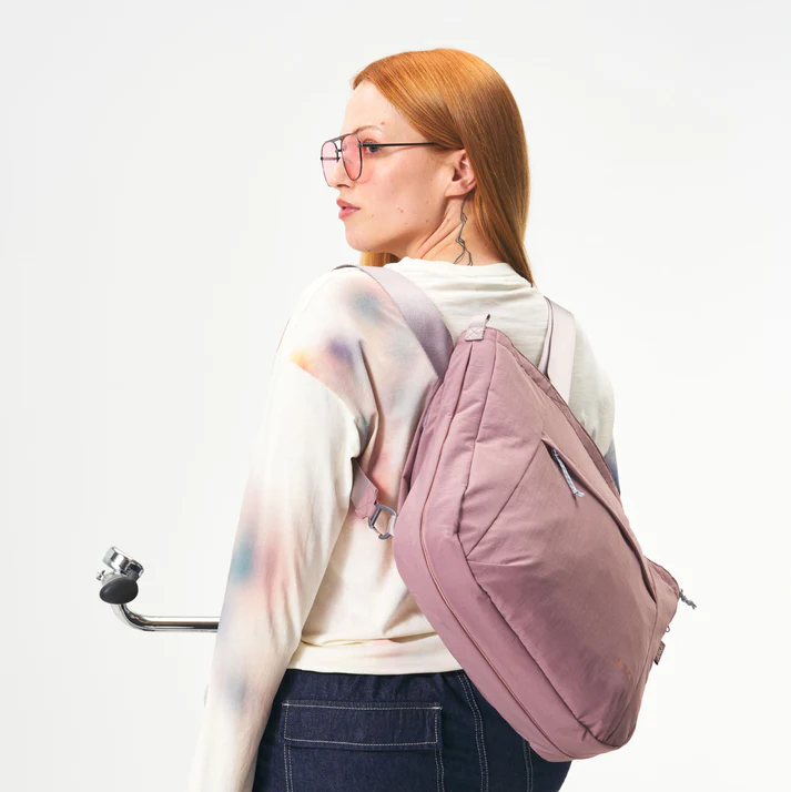 Eine Frau mit langen roten Haaren und Brille, die ein Batik-Sweatshirt und dunkle Jeans trägt, hat einen lilafarbenen Rucksack auf dem Rücken und hält einen Rollergriff vor einem schlichten weißen Hintergrund.