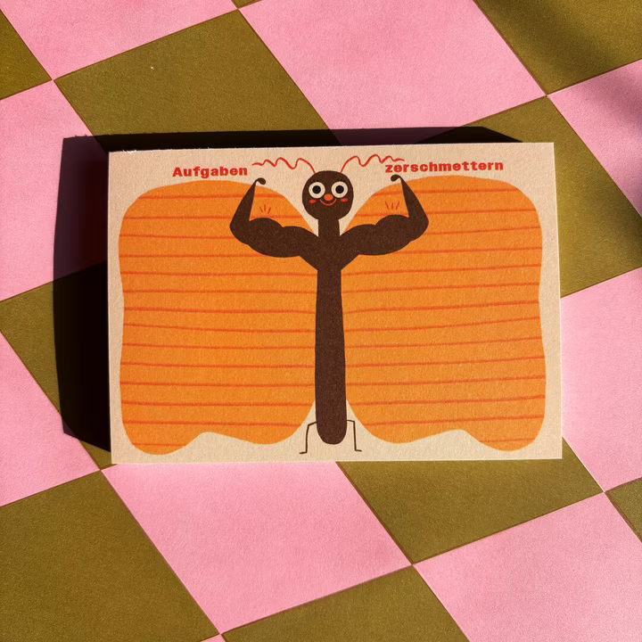 Ein Strichmännchen mit ausgestreckten Armen und großen Muskeln steht auf einem orangefarbenen Hintergrund; über seinen Armen stehen die Worte "Aufgaben" und "zerschmettern". Die Karte ist auf einer rosa und grün karierten Fläche platziert.