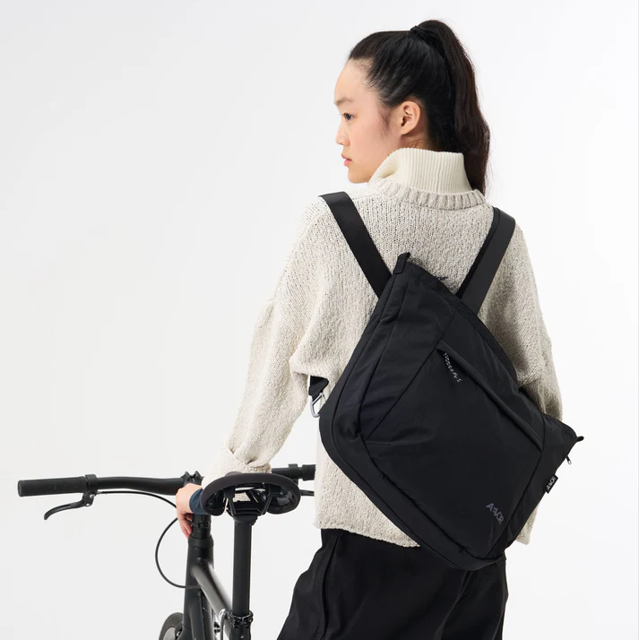 Eine Person mit einem hohen Pferdeschwanz, die einen cremefarbenen Pullover und eine schwarze Hose trägt, steht neben einem Fahrrad und trägt eine große schwarze Tasche auf dem Rücken. Der Hintergrund ist einfarbig weiß.