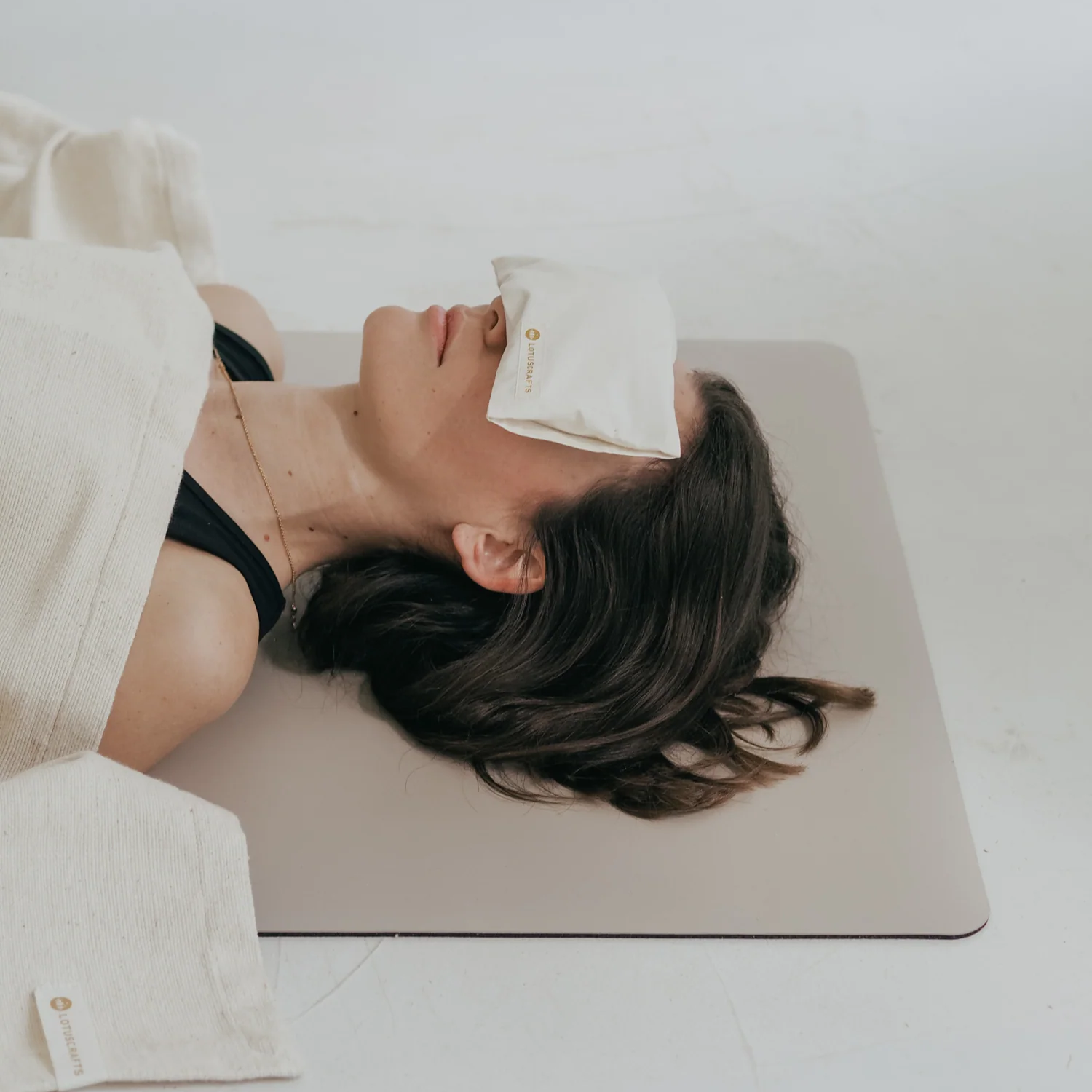 Eine Frau liegt auf dem Rücken auf einer Yogamatte, zugedeckt mit einer leichten Decke und einem kleinen Kissen über den Augen. Sie wirkt entspannt in einer ruhigen, minimalistischen Umgebung.