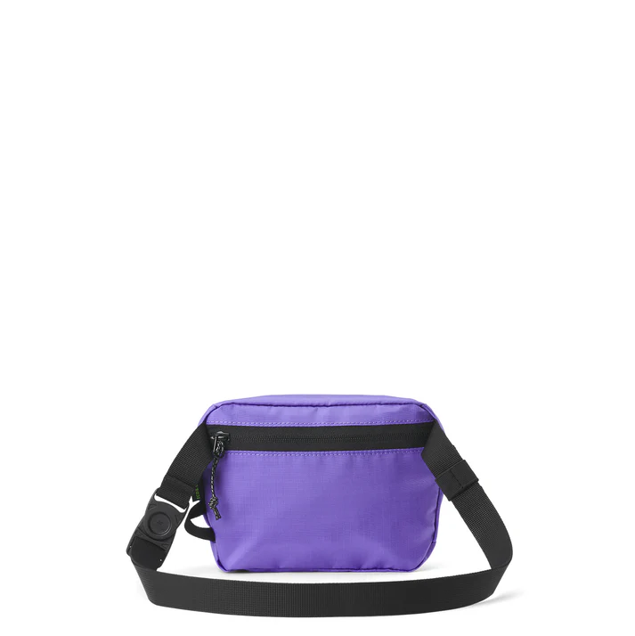 Eine kleine lilafarbene Umhängetasche mit schwarzem Reißverschluss und verstellbarem schwarzem Riemen, die vor einem schlichten weißen Hintergrund steht.