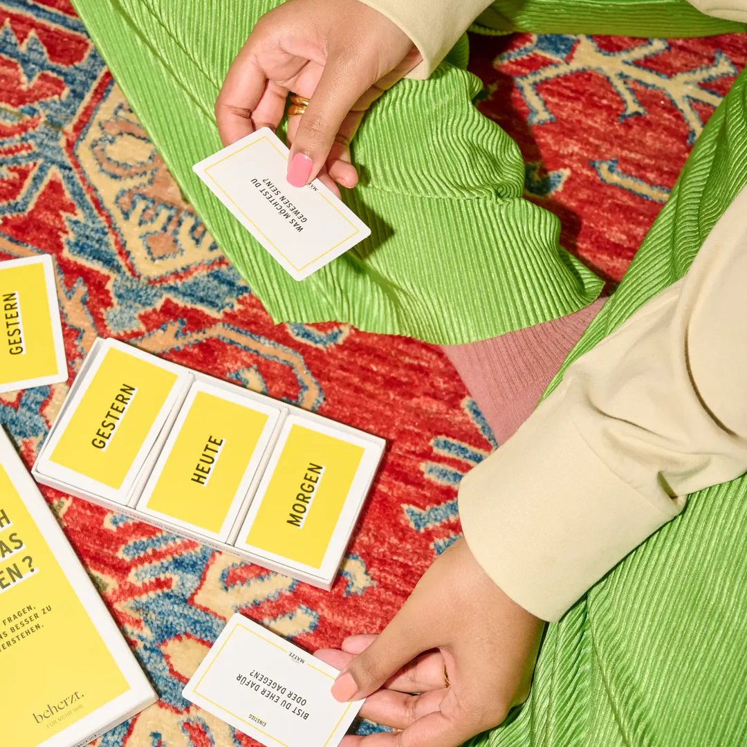 Eine Person in grüner Hose sitzt auf einem bunt gemusterten Teppich und hält Karten von einem gelb-weißen Kartenspiel in der Hand. Die Karten sind mit "GESTERN", "HEUTE" und "MORGEN" beschriftet, was auf Deutsch "gestern", "heute" und "morgen" bedeutet.