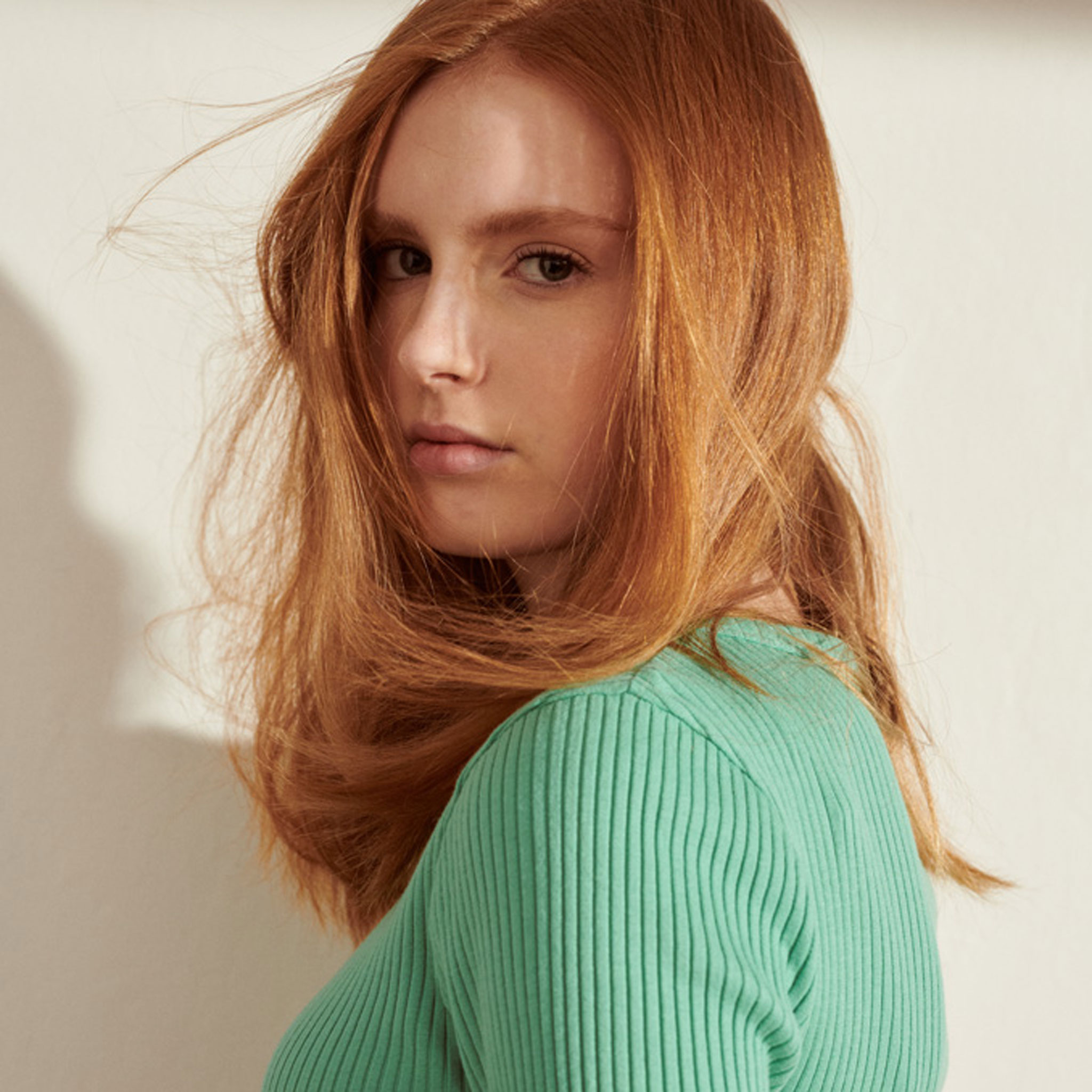 Eine Frau mit langen roten Haaren trägt einen grünen Pullover und schaut zur Seite, während sie im Licht steht.