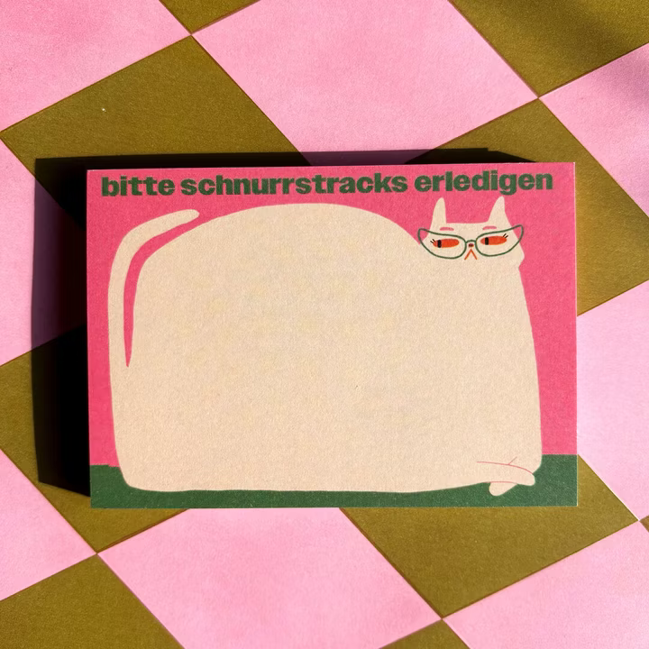 Ein Notizblock mit einer großen Cartoon-Katze, die eine Brille trägt, auf einem rosa Hintergrund. Der deutsche Text "bitte schnurrstracks erledigen" steht oben auf dem Notizblock. Der Block ist auf einer rosa und braun karierten Oberfläche.